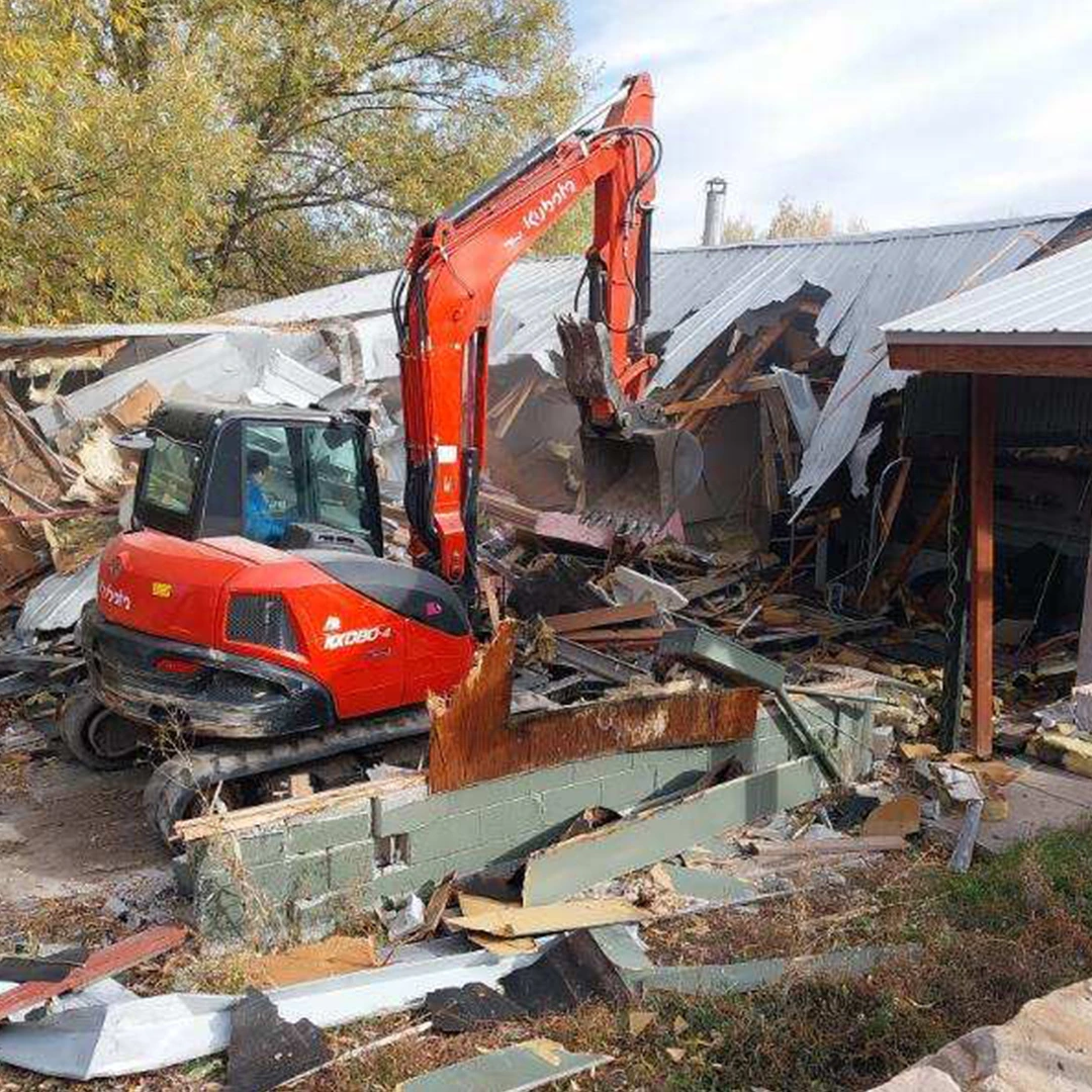 A red excavator demolishes a building, surrounded by debris including wooden beams and metal scraps on the ground.