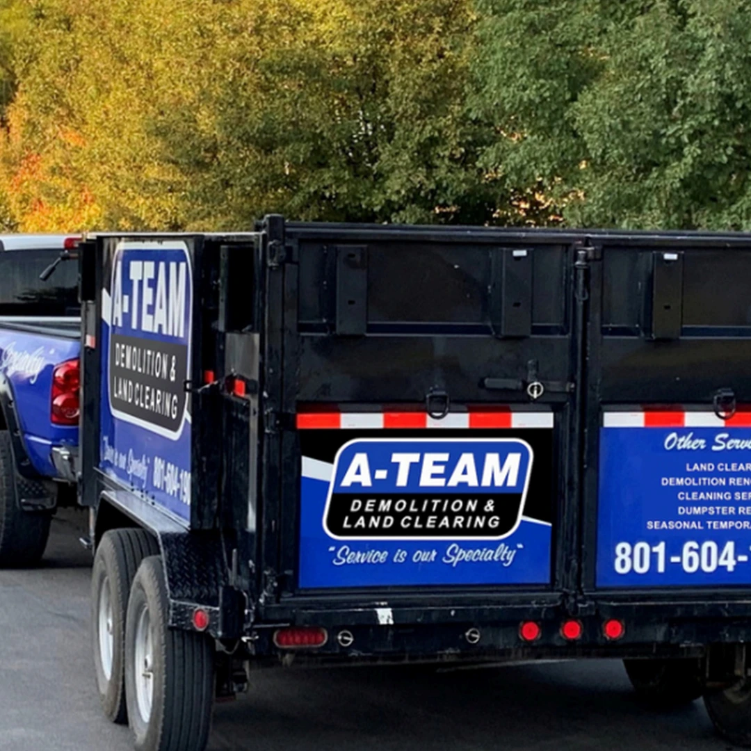 A close-up of an A-Team Demolition & Land Clearing trailer parked outdoors, featuring a blue and black logo with contact info.